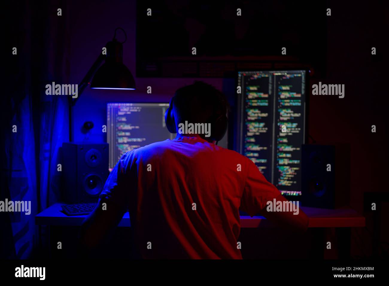 A man sits at a computer in a room at a table at night with blue lighting and programs Stock Photo