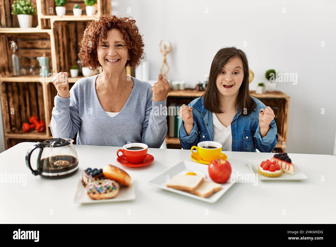 Family of mother and down syndrome daughter sitting at home eating breakfast celebrating surprised and amazed for success with arms raised and open ey Stock Photo