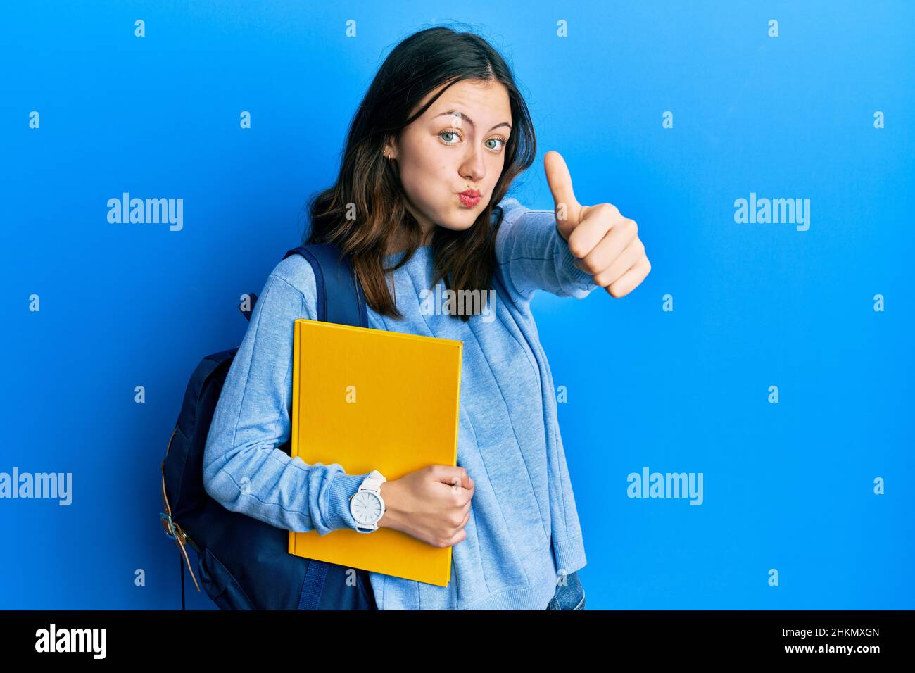 Young brunette student woman wearing student backpack doing thumbs up gesture puffing cheeks with funny face. mouth inflated with air, catching air. Stock Photo