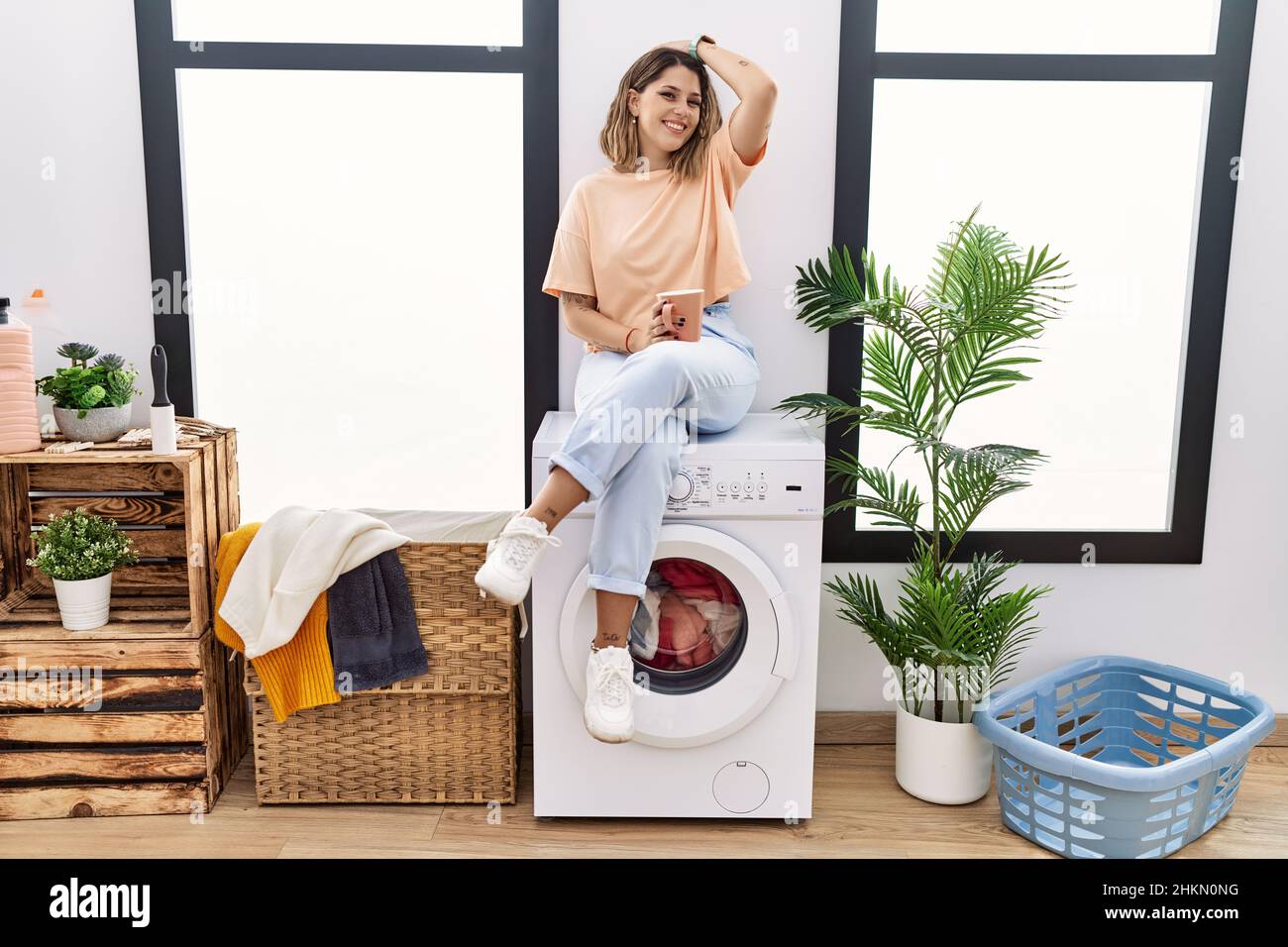 Young hispanic woman drinking coffee waiting for washing machine at laundry room smiling confident touching hair with hand up gesture, posing attracti Stock Photo