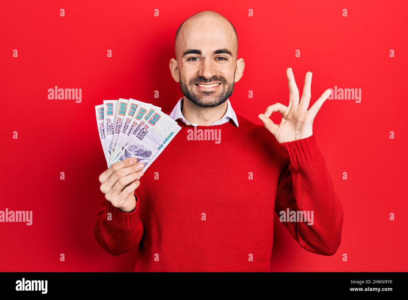 Young bald man holding egyptian pounds banknotes doing ok sign with fingers, smiling friendly gesturing excellent symbol Stock Photo