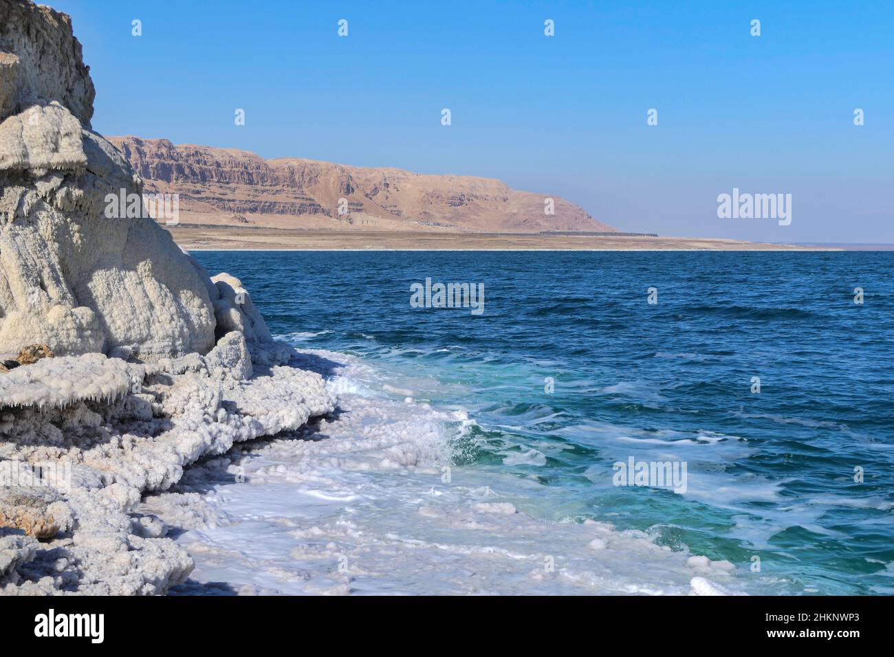 View of the mountains across the waters of the Dead Sea from the shore covered with salt formations. Stock Photo