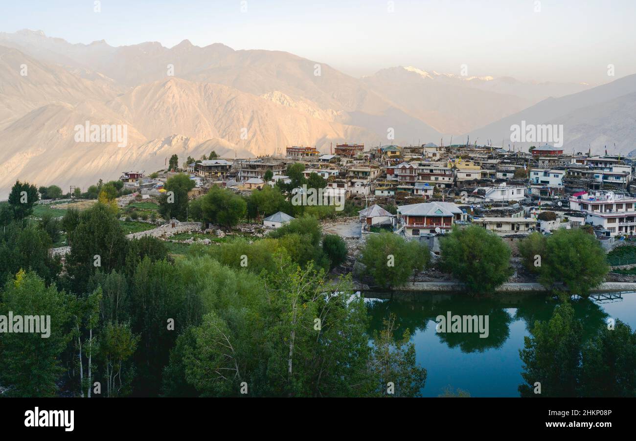 Elevated view of small town  with buildings flanked by lake, trees, and the rugged slopes and peaks of the Himalayas at sunrise in Nako, India. Stock Photo