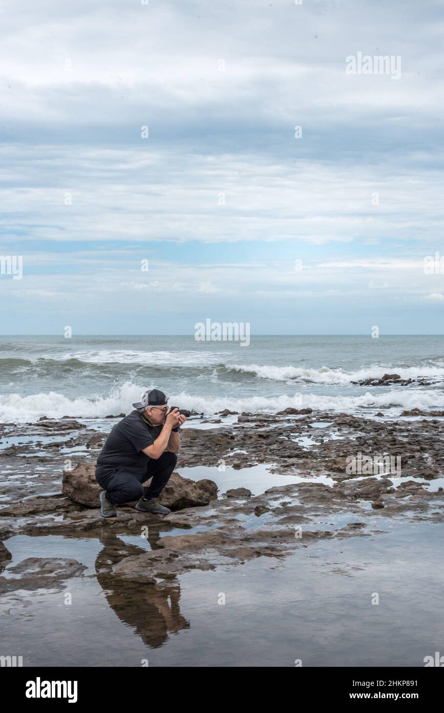 Adult man with glasses kneeling on the rocks taking a photograph. The ...