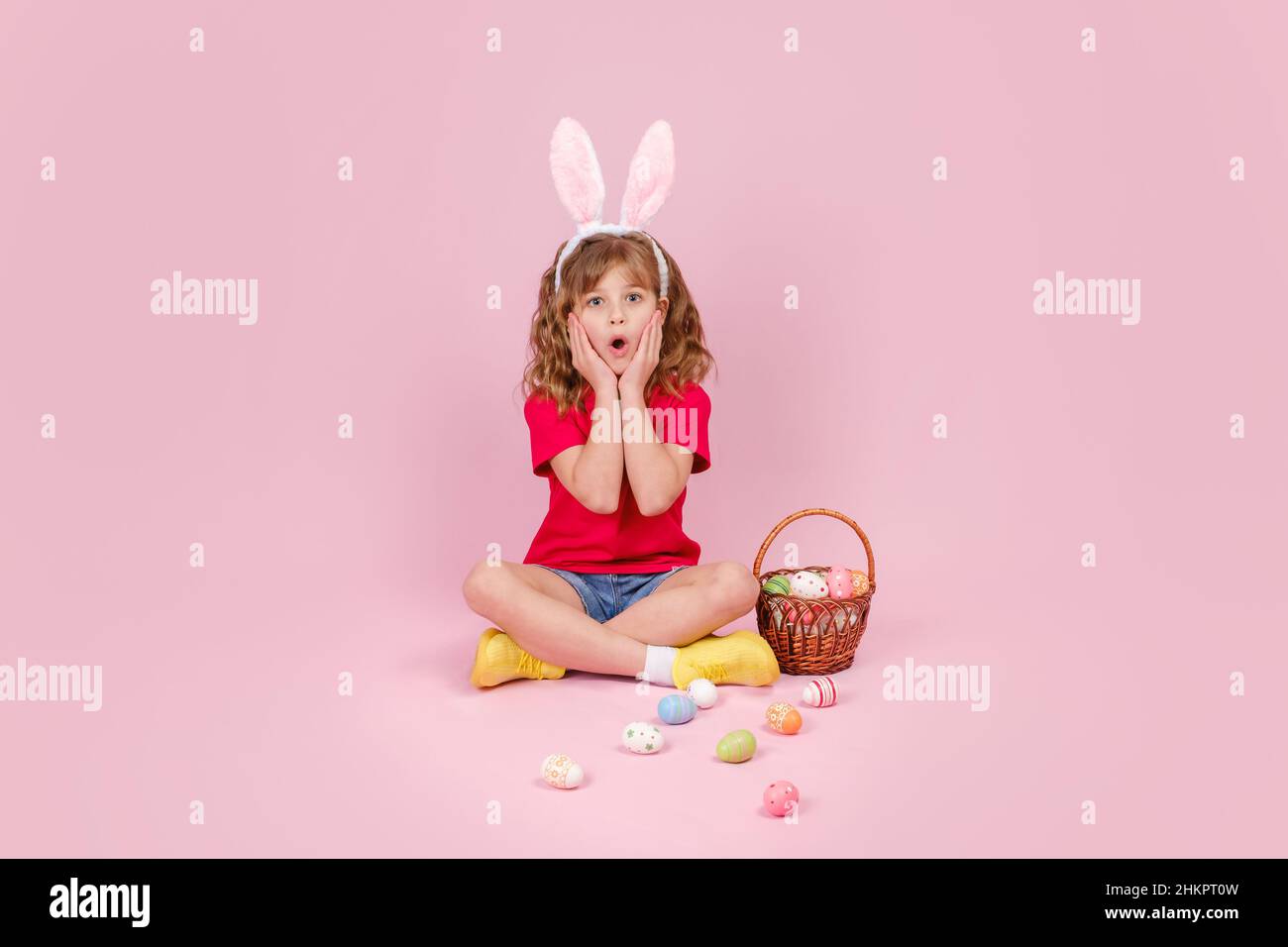 Girl in Easter bunny ears sitting on the floor and covering eye with egg Stock Photo
