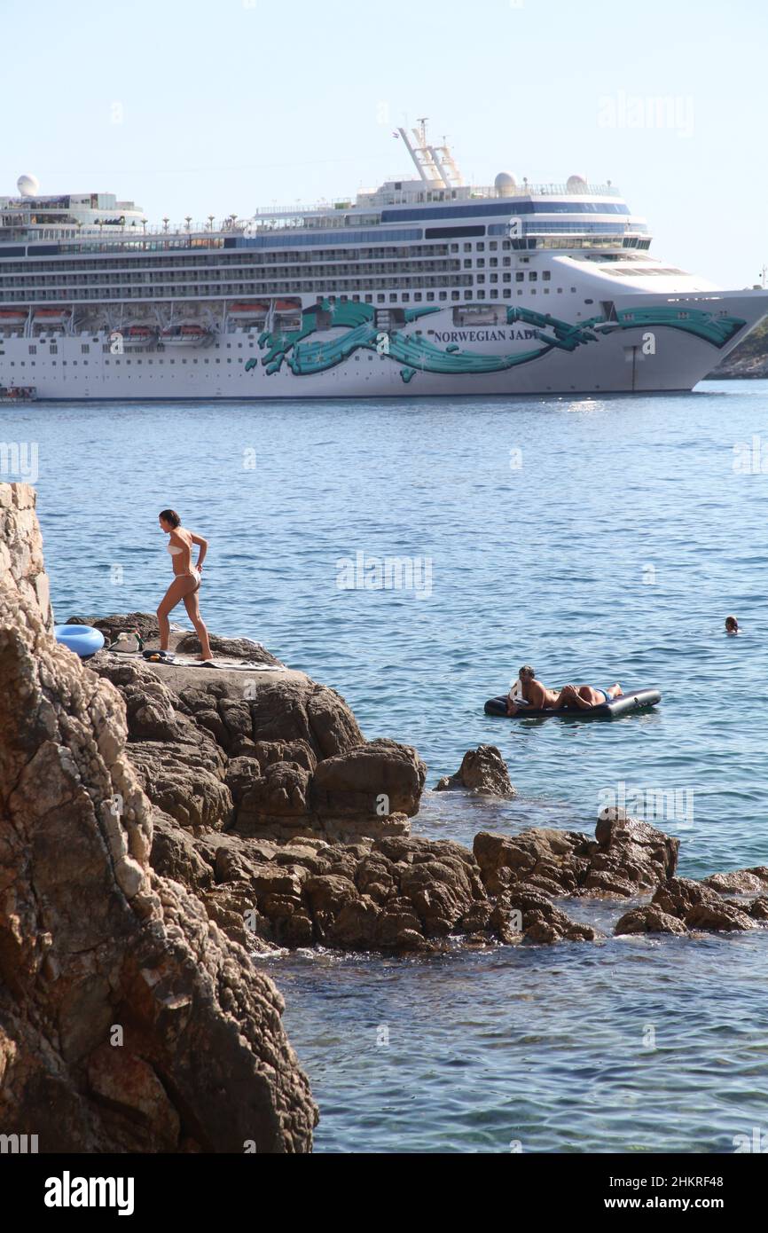 Young lady tourist steps on rock from water fitting her panties as Norwegian Jade cruise ship passes by St Jacob beach near Dubrovnik, Adriatic shore Stock Photo
