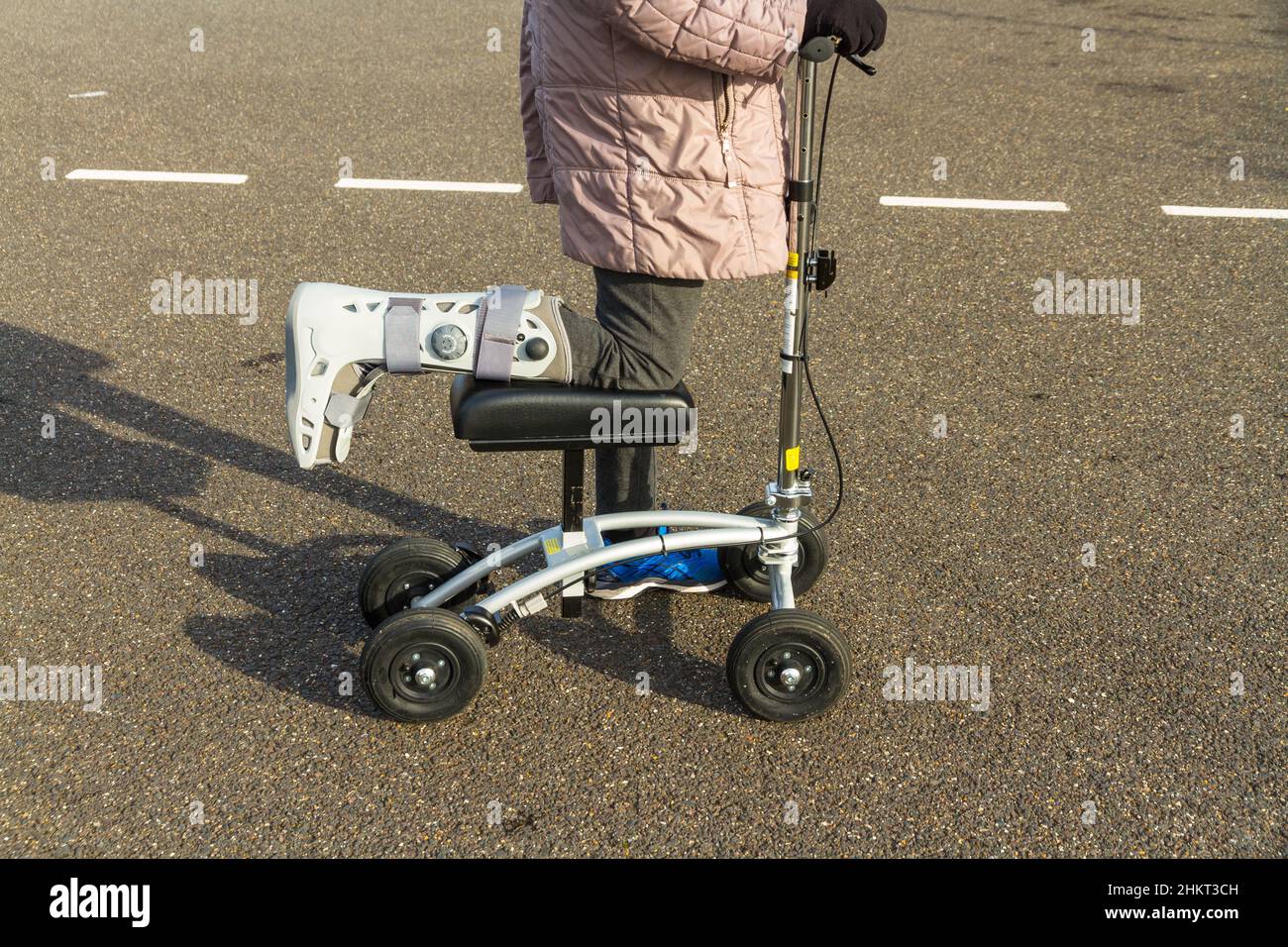 Knee scooter or walker being used by woman with leg foot in surgical boot on sunny winter day, landscape. Stock Photo