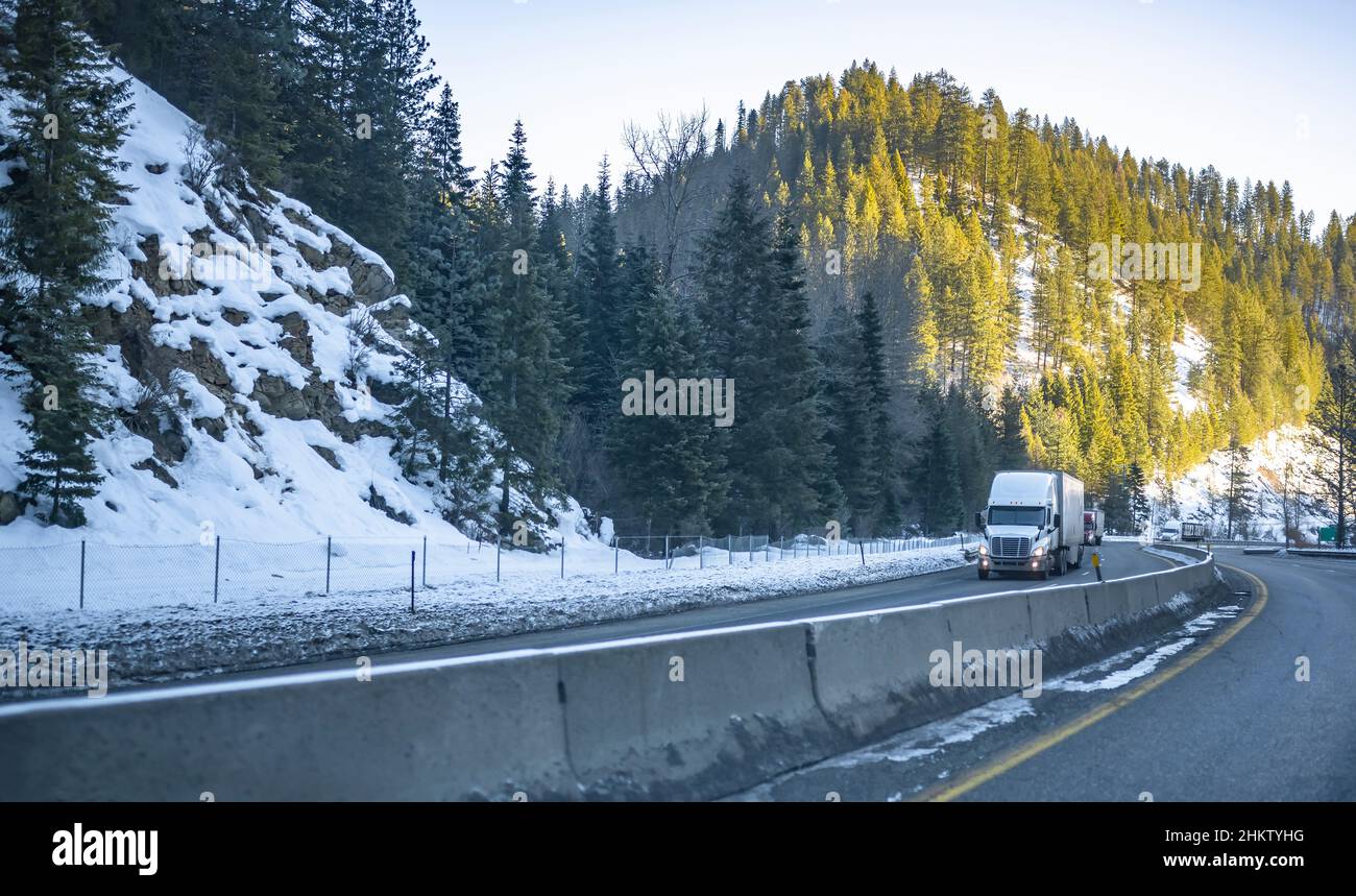 Big rig semi trucks tractors transporting commercial cargo in semi trailers driving in convoy on winding winter slippery highway road with snow and ic Stock Photo