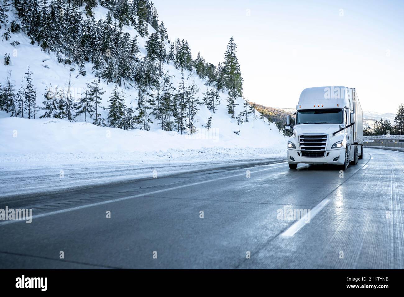 White big rig industrial semi truck transporting cargo in dry van semi trailer running on the winding winter dangerous slippery road with snow and ice Stock Photo
