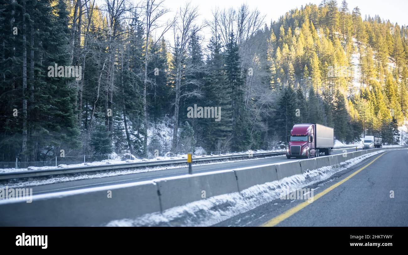 Convoy of industrial big rigs semi trucks transporting cargo in semi trailers running on the winding winter dangerous slippery road with snow and ice Stock Photo