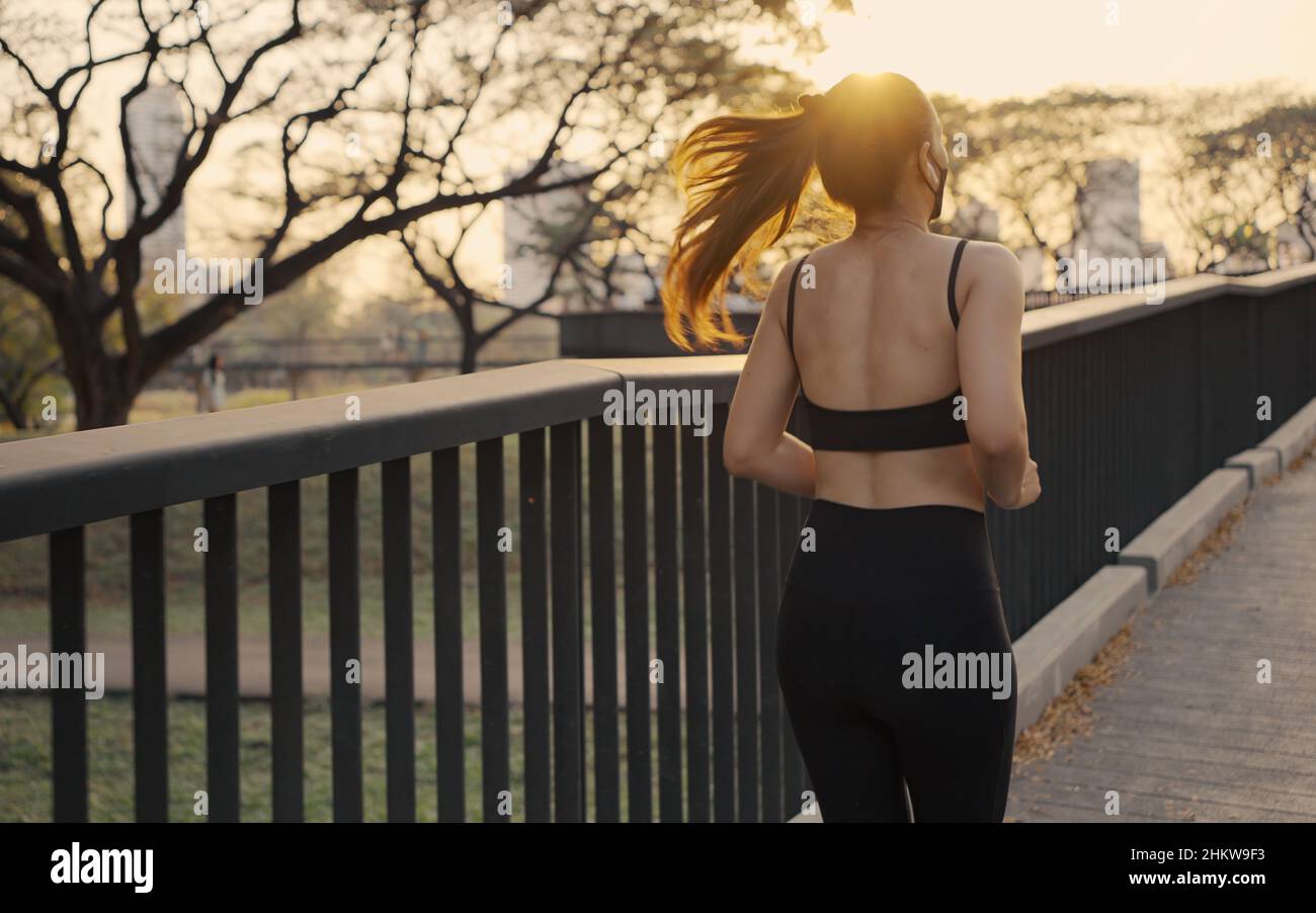Woman in sportswear wearing a protective mask jogging exercise on the bridge in the city at sunset. Stock Photo