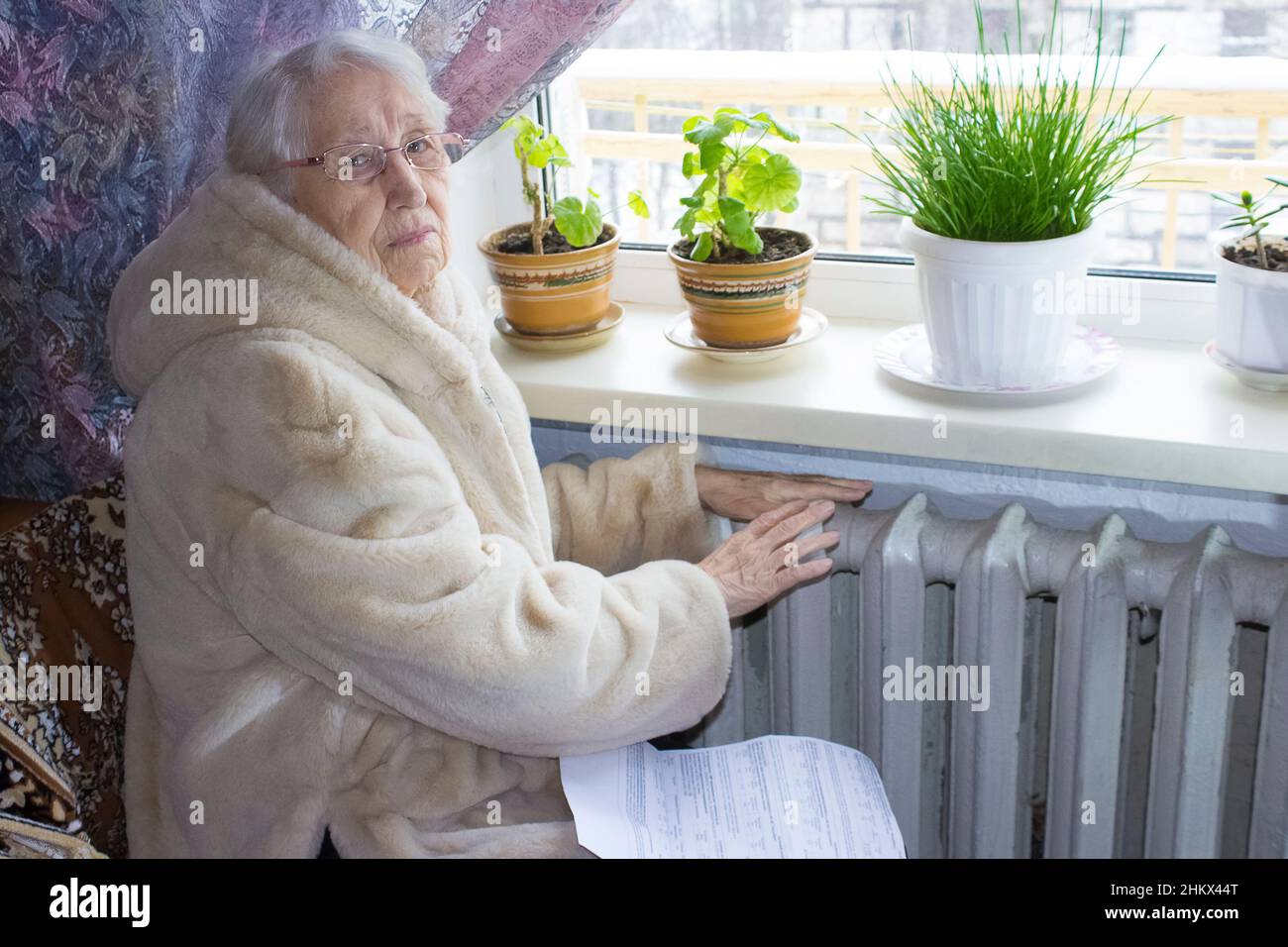 The senior woman holding gas bill in front of heating radiator. Payment for heating in winter. Stock Photo
