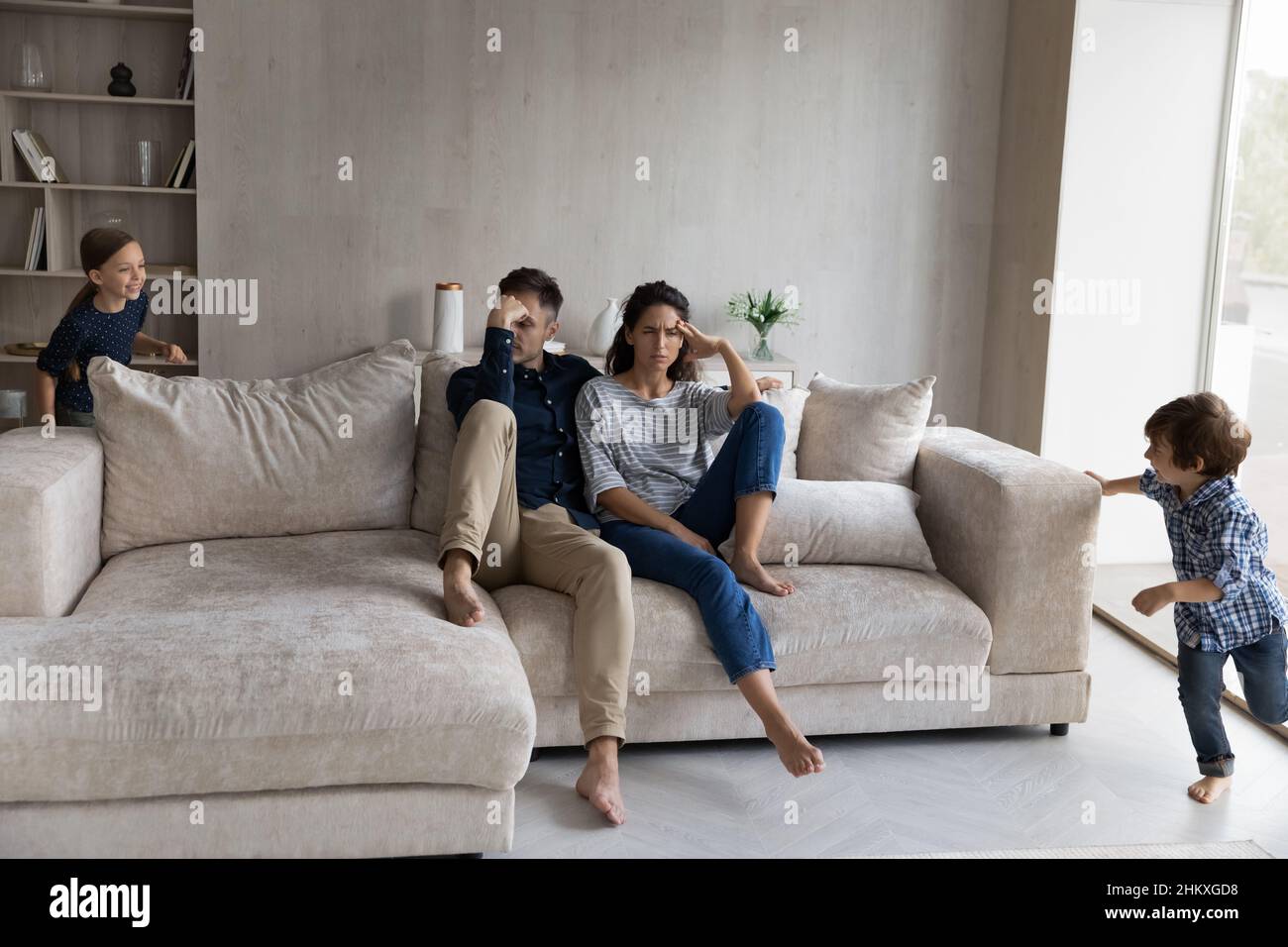 Tired parents sitting on couch while noisy kids running around Stock Photo