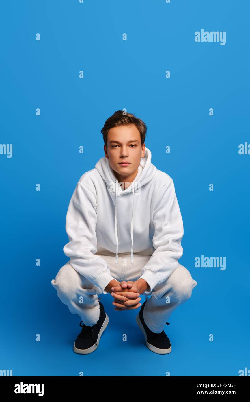 Attractive man wearing white sweatshirt and sweatpants is squatting over blue studio background Stock Photo