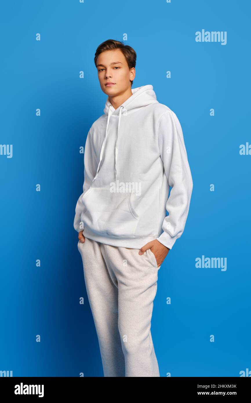 Young man wearing white sweatshirt and sweatpants standing over blue studio background with hands in pockets Stock Photo