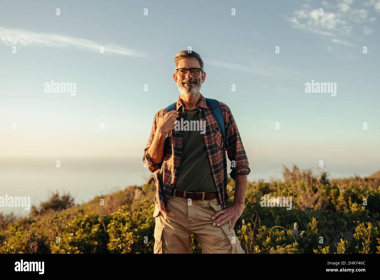 Mature hiker smiling at the camera at sunset. Happy mature hiker standing on top of a hill while carrying a backpack. Adventurous male backpacker enjo Stock Photo