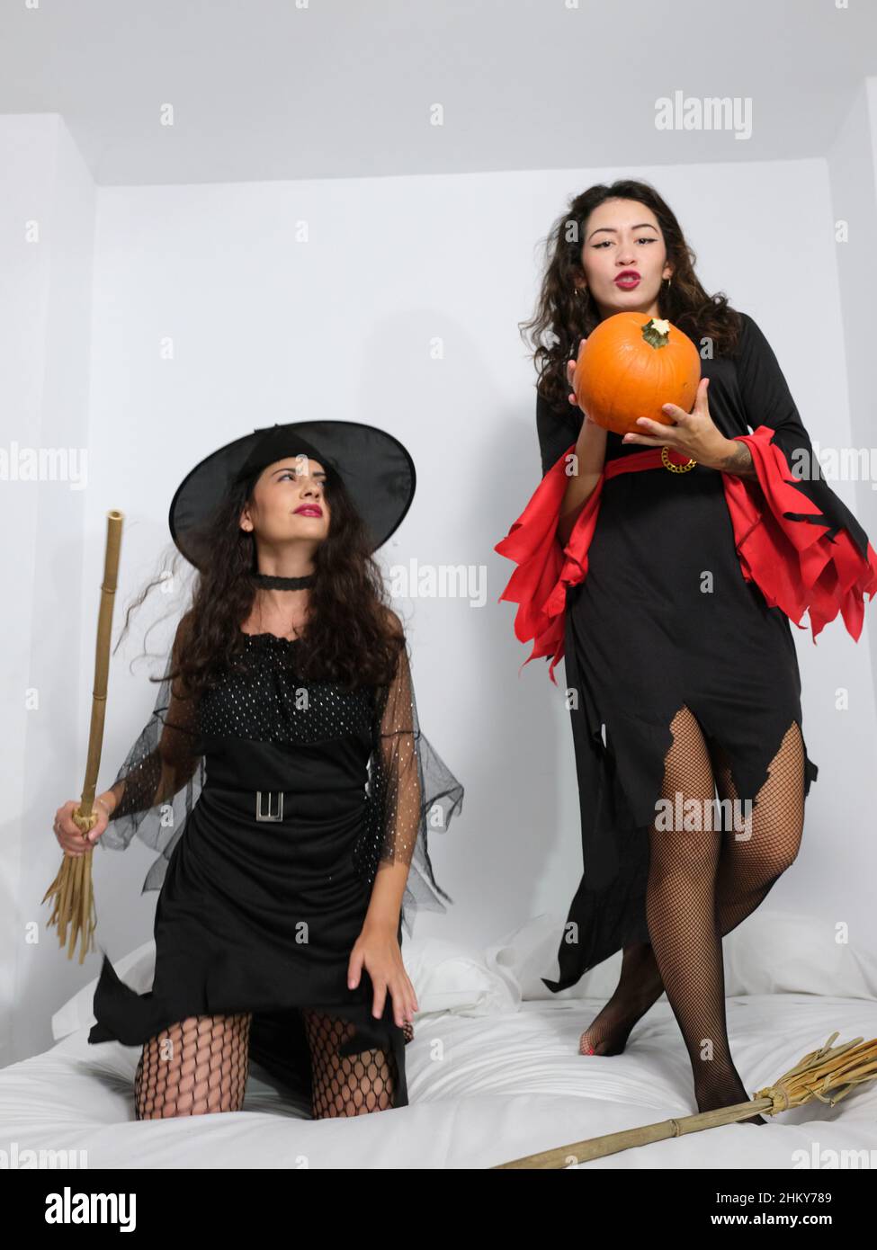 Two young latinas dressed as witches in halloween costumes having fun on the bedroom bed with a pumpkin Stock Photo