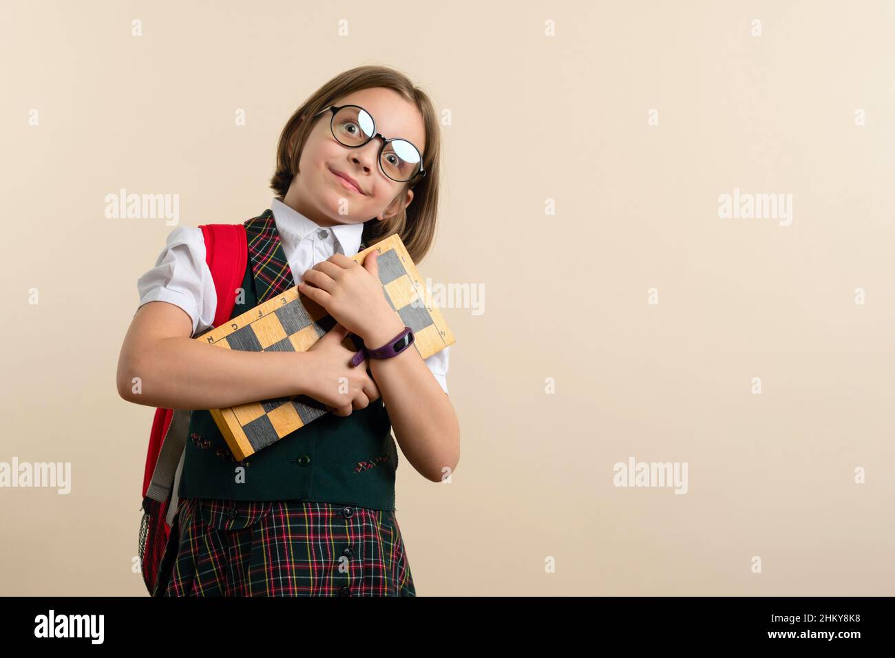 Excited Nerd Little Girl 9s wearing school uniform and glasses hug chessboard. Child with educational game. Learn to play chess. Child chess player. Stock Photo