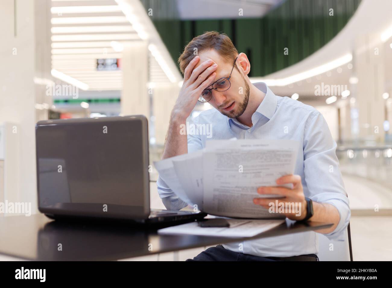 Young puzzled businessman working in empty food court in mall with documents and laptop. Filing tax returns work with contracts Stock Photo