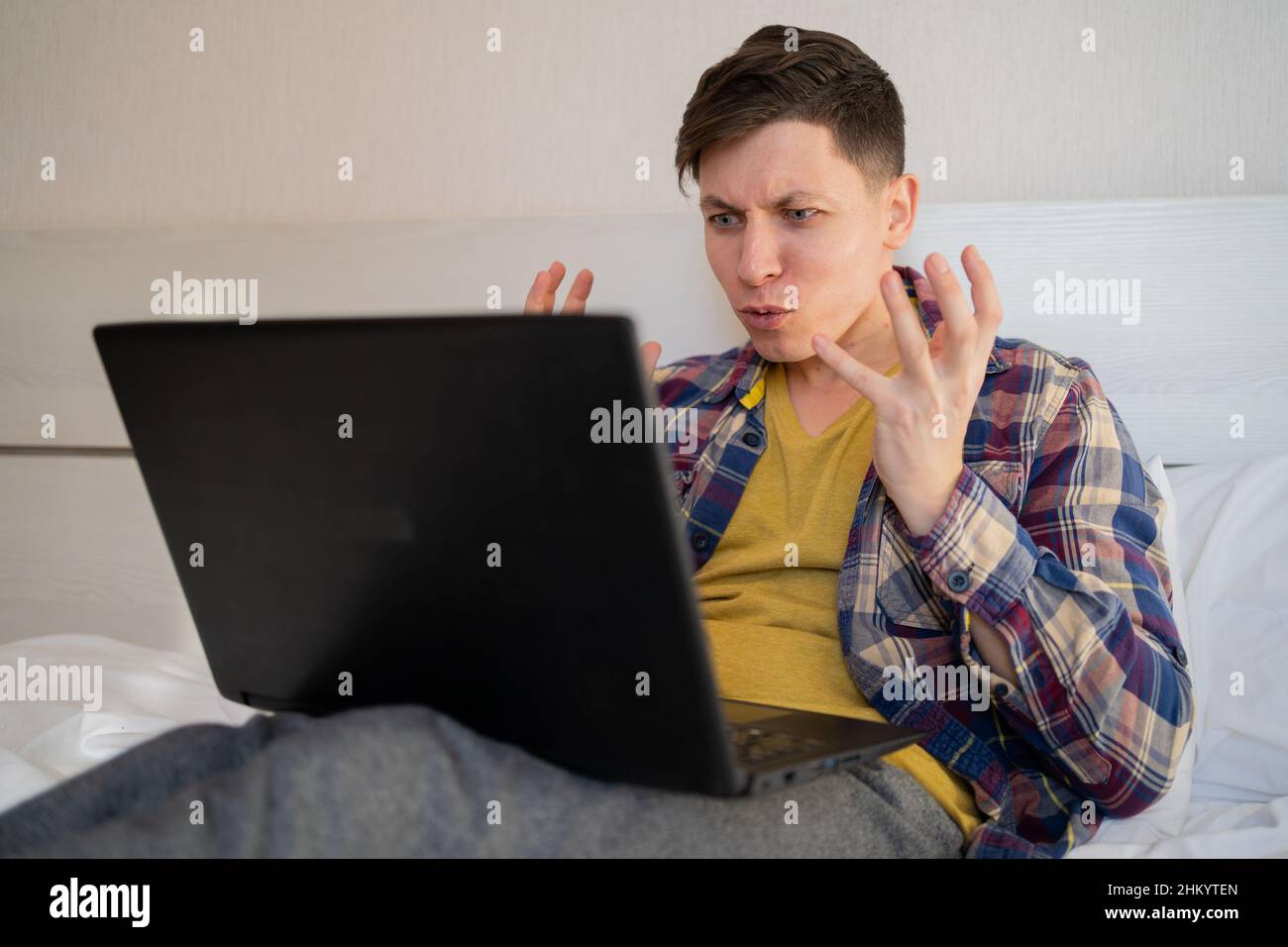 Irritated man sitting on bed looking at laptop screen, arms