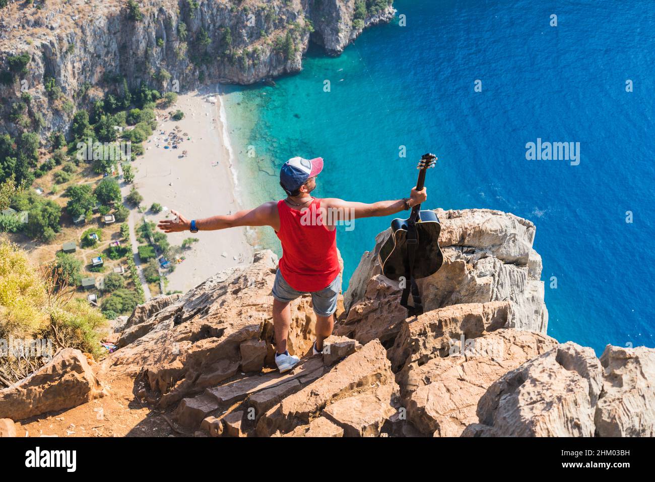 Musician man standing and holding guitar with open arms at the edge of a cliff. Butterfly valley in the background. Stock Photo