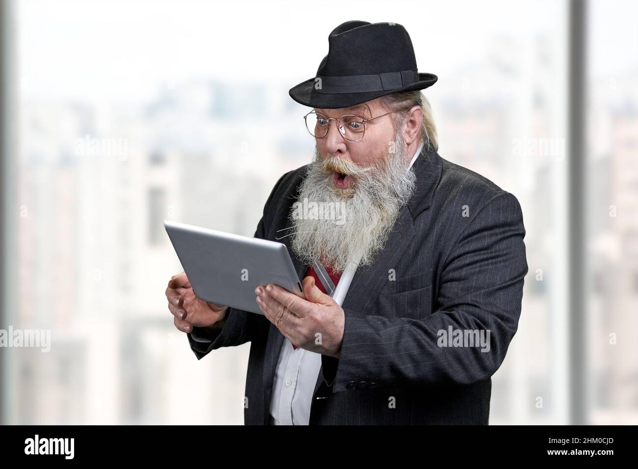Funny shocked senior businessman using tablet pc. Blurred office window background. Stock Photo