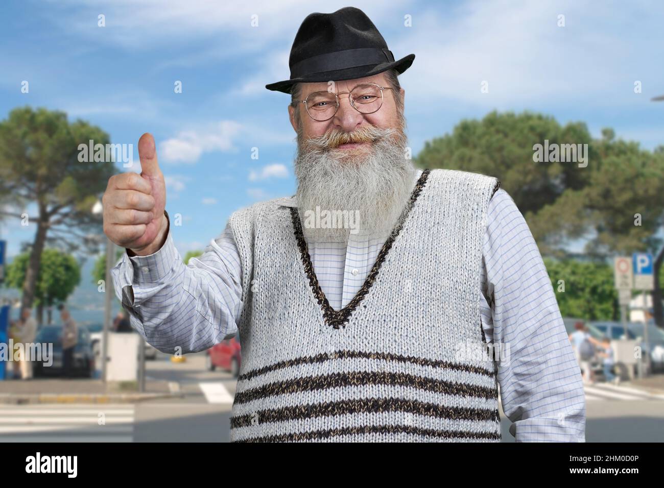 Old bearded man showing thumb up standing outdoors. Happy grandfather looking at camera and gesturing thumb up. Stock Photo