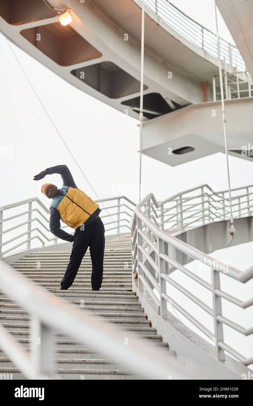 Young African American sportsman in tracksuit and sleeveless jacket practicing side bends on staircase in urban environment Stock Photo