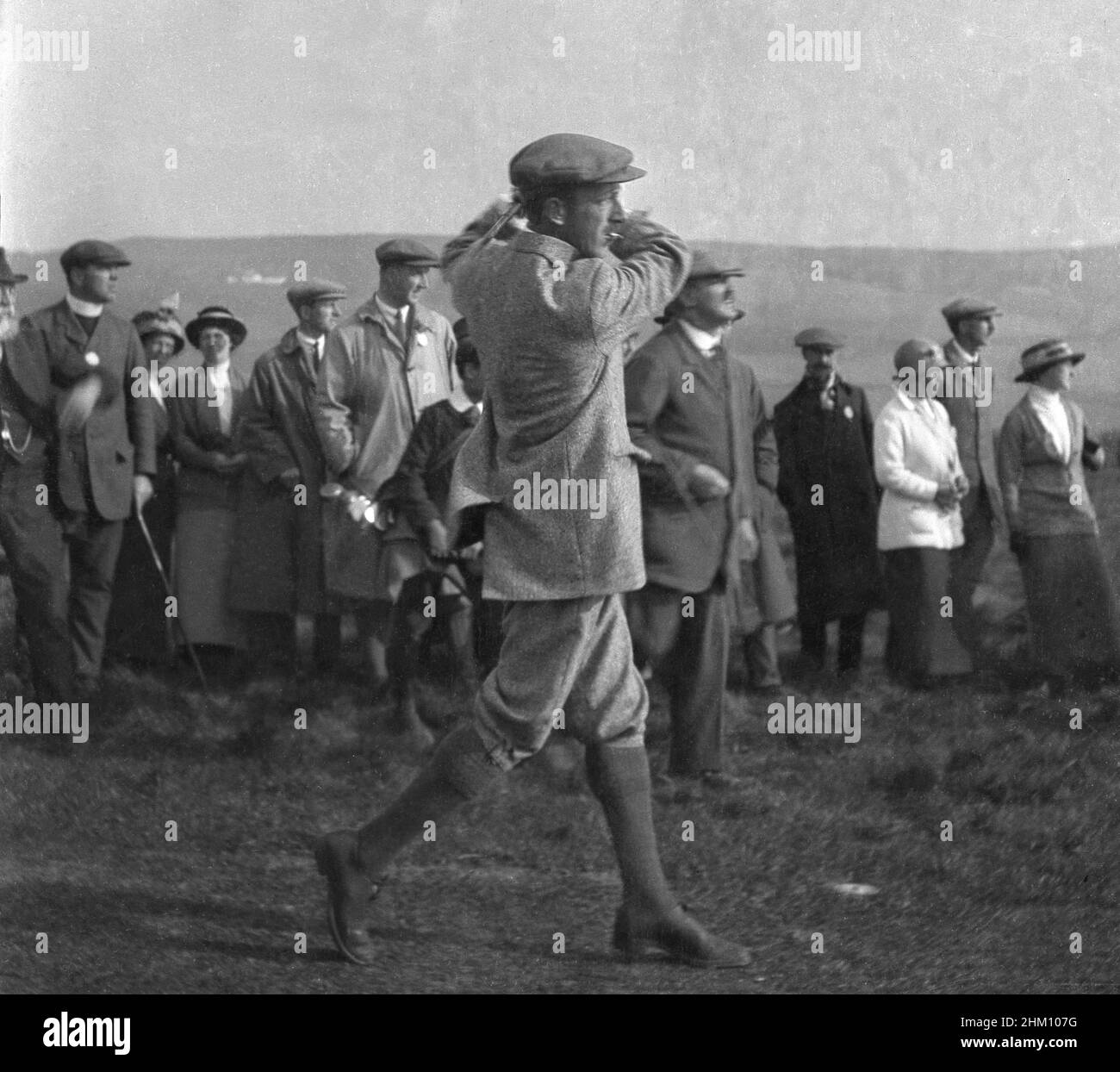 circa 1910, historical, a professional male golfer - possibly Harry Vardon - in a jacket, plus-twos and a cloth cap and cigarette in his mouth hiting a shot, watched by a small group of spectators in the formal clothing of the time. His playing partner, J H. Taylor, co-founder of the British PGA in 1901 and a five-time winner of the British Open Championship was a golfing pioneer of the era. Stock Photo