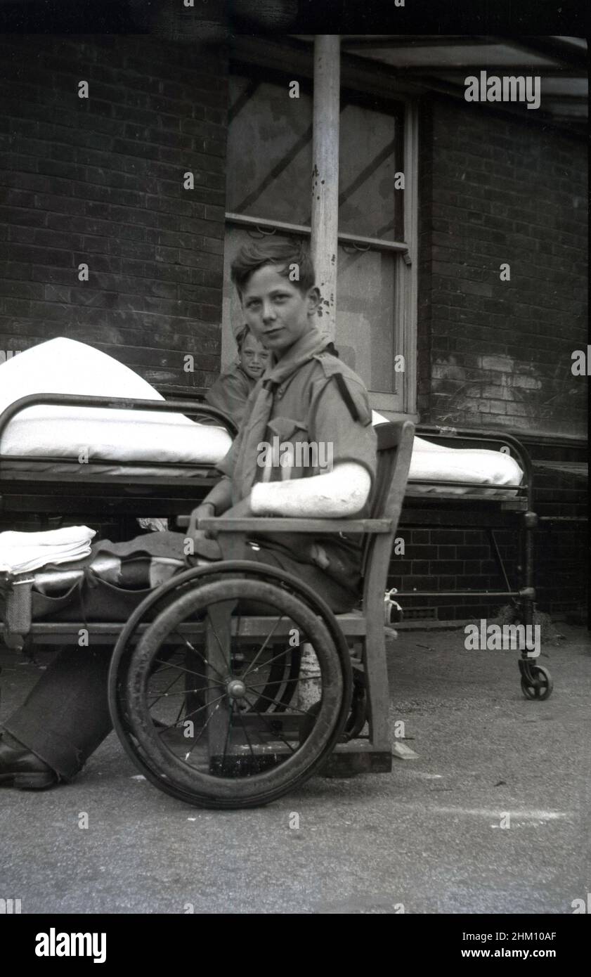 1942, outdoors, beside his metal bed, a boy scout sitting in a wheelchair of the era, leg outstretched in a casing and one of his arms in a plaster cast, Queen Mary's Hospital in Carshalton, Surrey, England, UK. Opened in 1908 as the Southern Hospital, in 1909 the poor and sick children of London, many who had TB went there and it was renamed the Children's Infirmary. The young patients were housed in single storey ward blocks.  In 1915 it was renamed Queen Mary's Hospital for Children when she became its patron. During WW2, many scouts who were injured during the London Blitz were sent there. Stock Photo