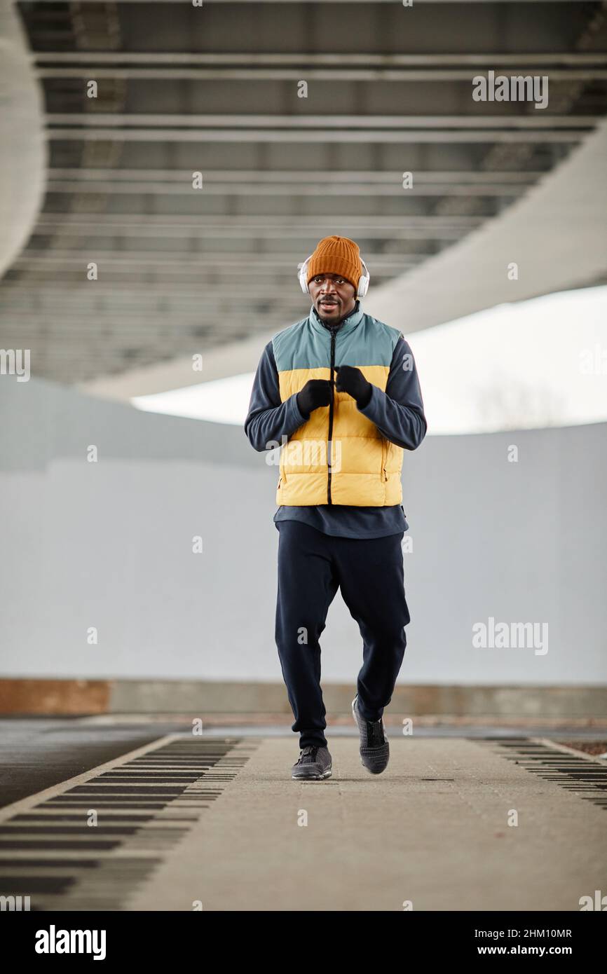 Young contemporary active man in headphones, warm tracksuit, hat and sleeveless jacket jogging along track in the morning Stock Photo