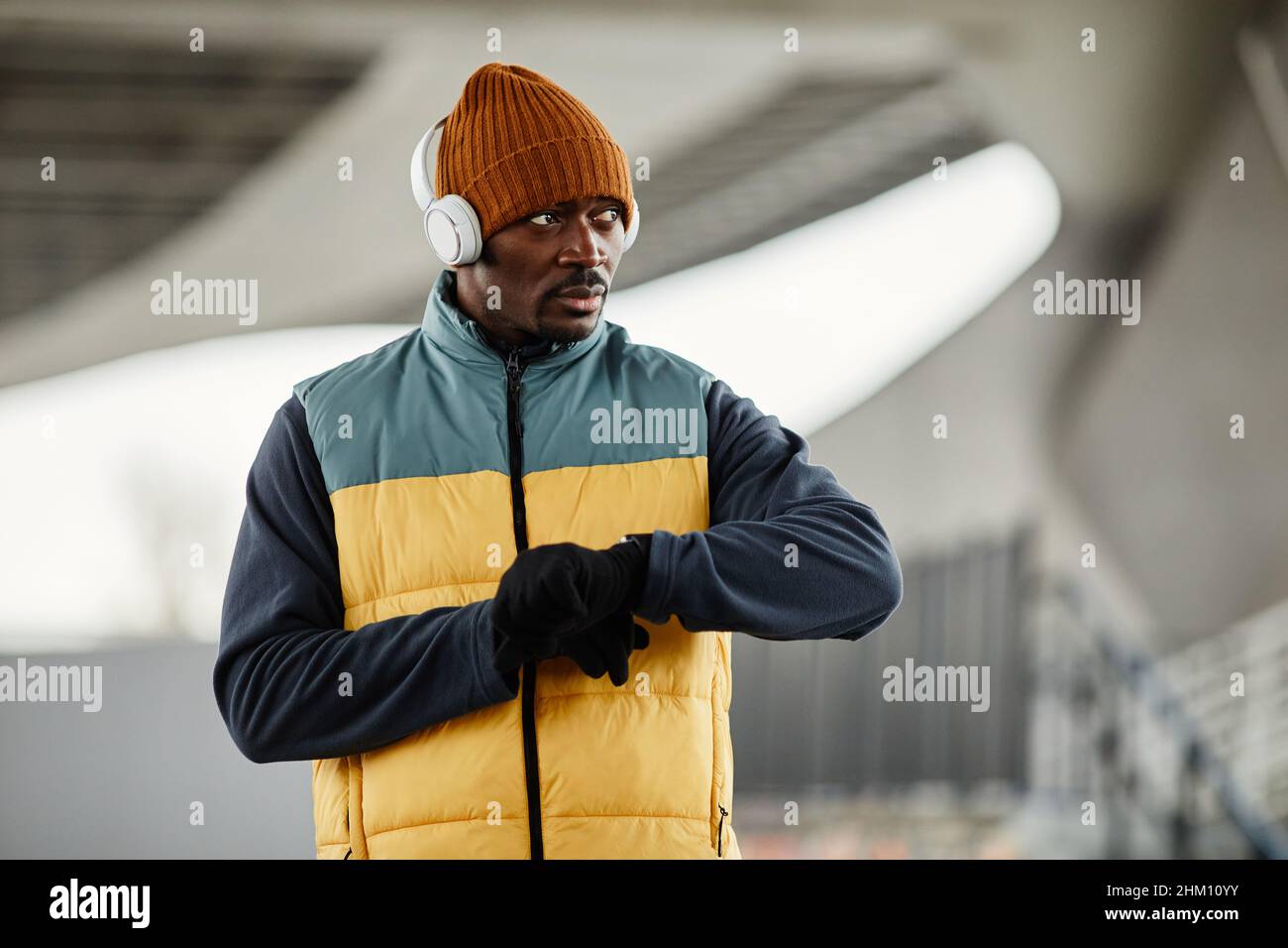 Contemporary young black man in headphones over beanie hat, sleeveless jacket on tracksuit and gloves Stock Photo