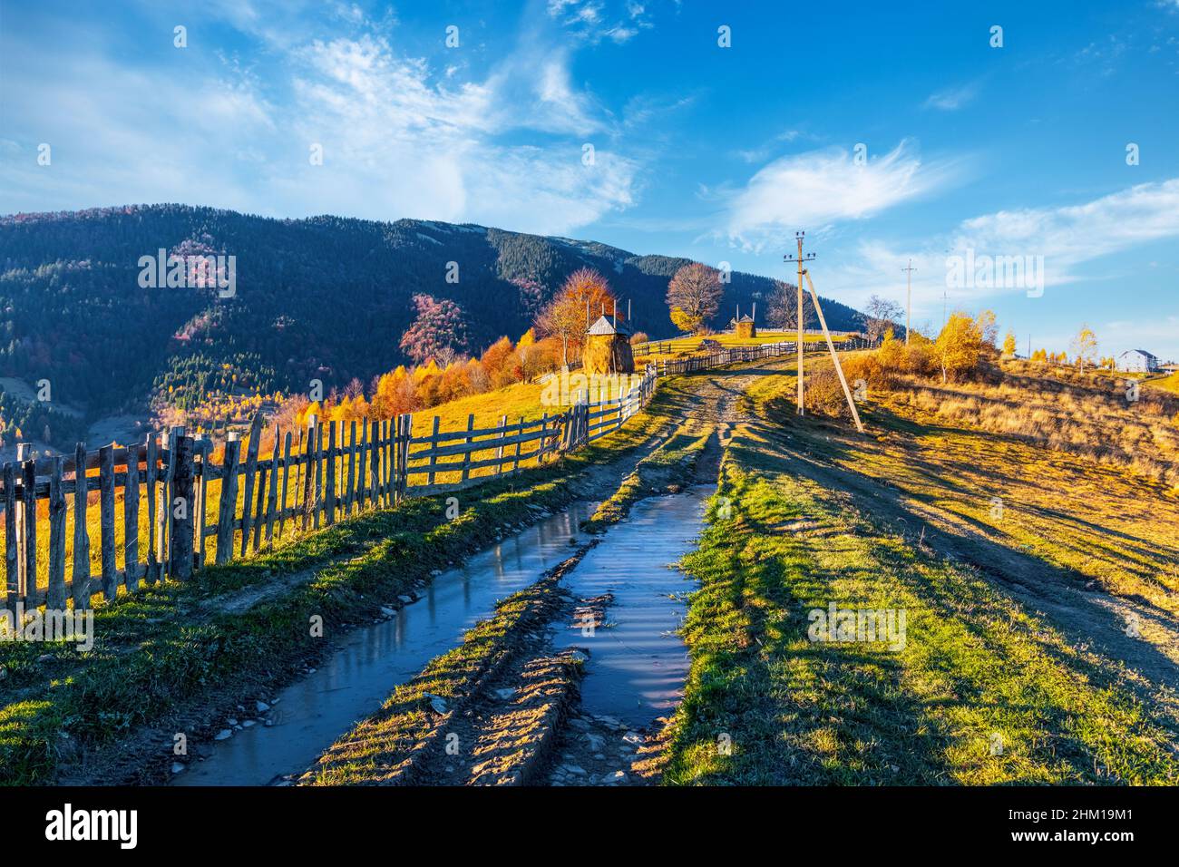 Ground road with dirty puddles after rain runs along wooden fence near highland village against forestry mountains on sunny autumn day Stock Photo