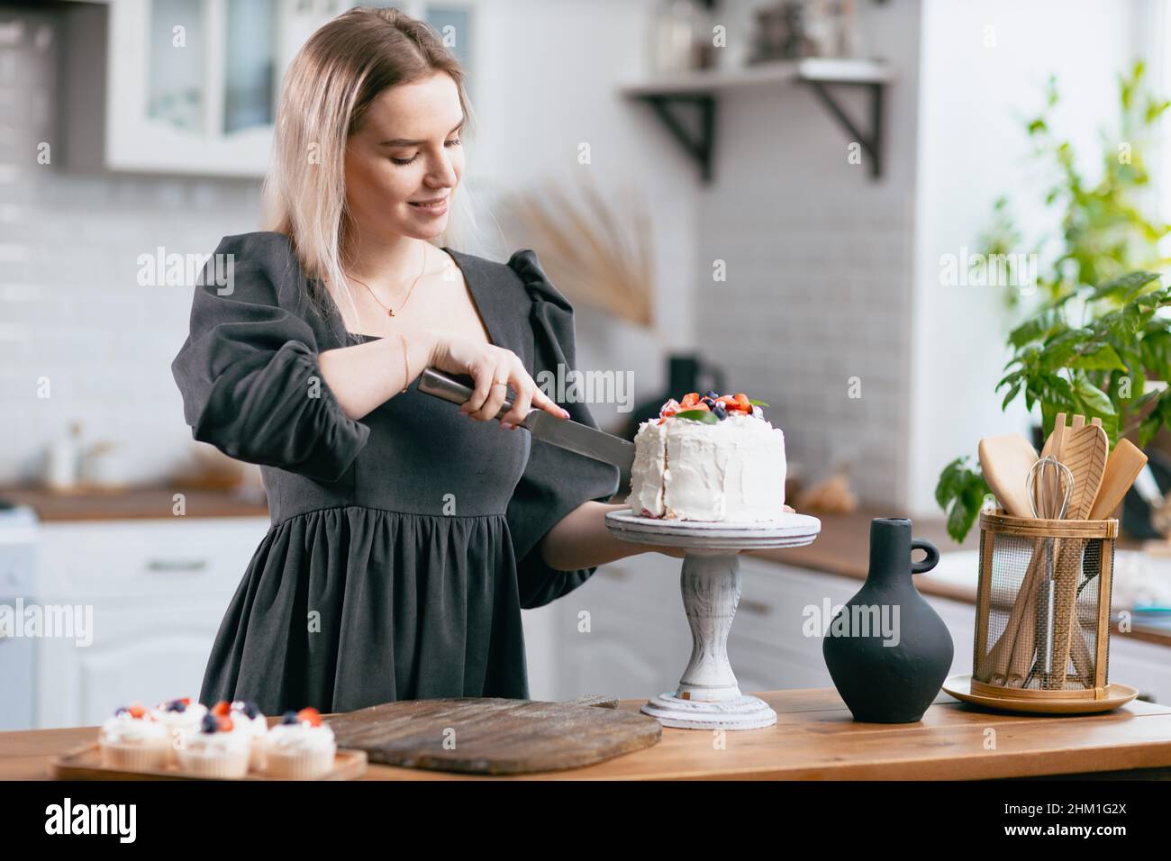 Pastry chef confectioner young caucasian woman in gray dress with knife cut slice cake on kitchen table. Cakes cupcakes and sweet dessert Scandinavian style Stock Photo