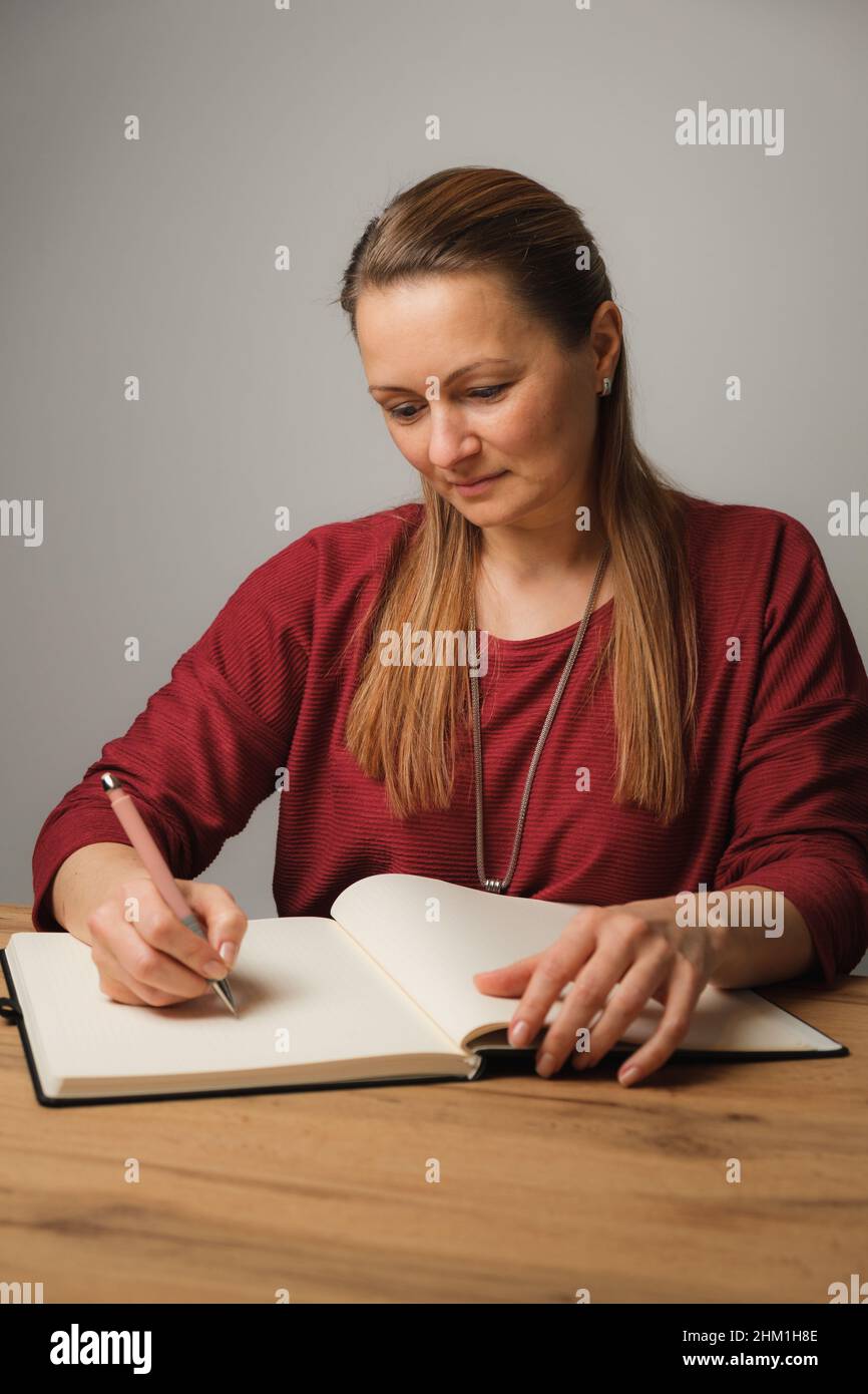 Young woman with long blond hair and a red pullover writes in a book while sitting at a wooden table Stock Photo