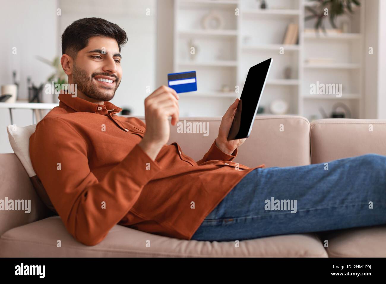 Smiling Arab guy using tablet and credit card at home Stock Photo