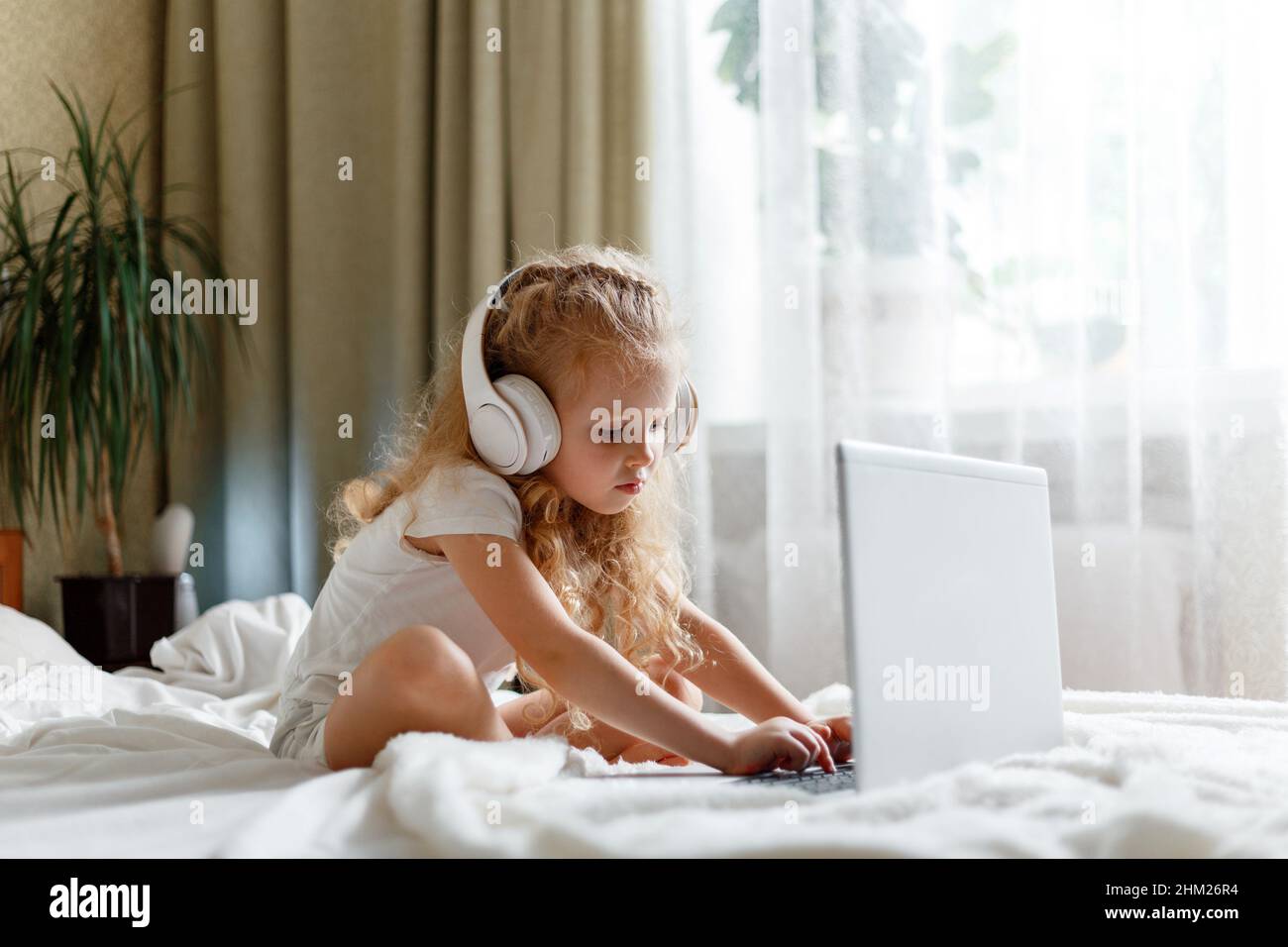 Preschooler girl in headphones use laptop for remote learning in kindergarten. Smart calm attentive girl child using laptop for watching video doing Stock Photo