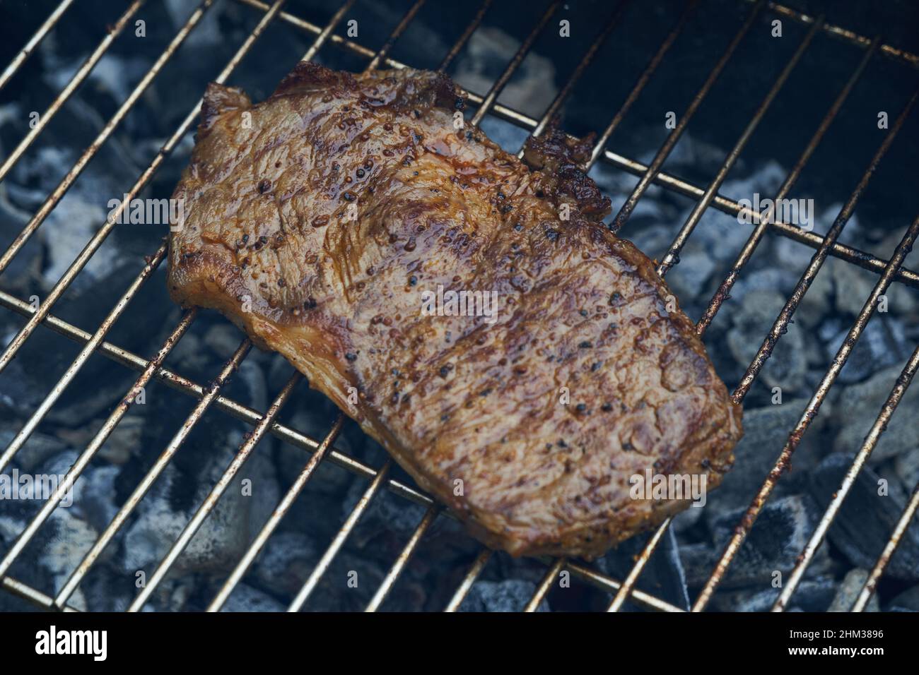 1 steak (rump steak) of beef lies on a stainless steel grill. Glowing coals roast the delicacy.Slight smoke development. View from above. Stock Photo