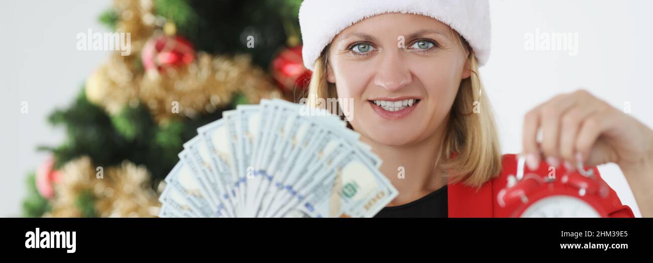 Woman in red santa hat holding money and alarm clock in hands near new year tree Stock Photo