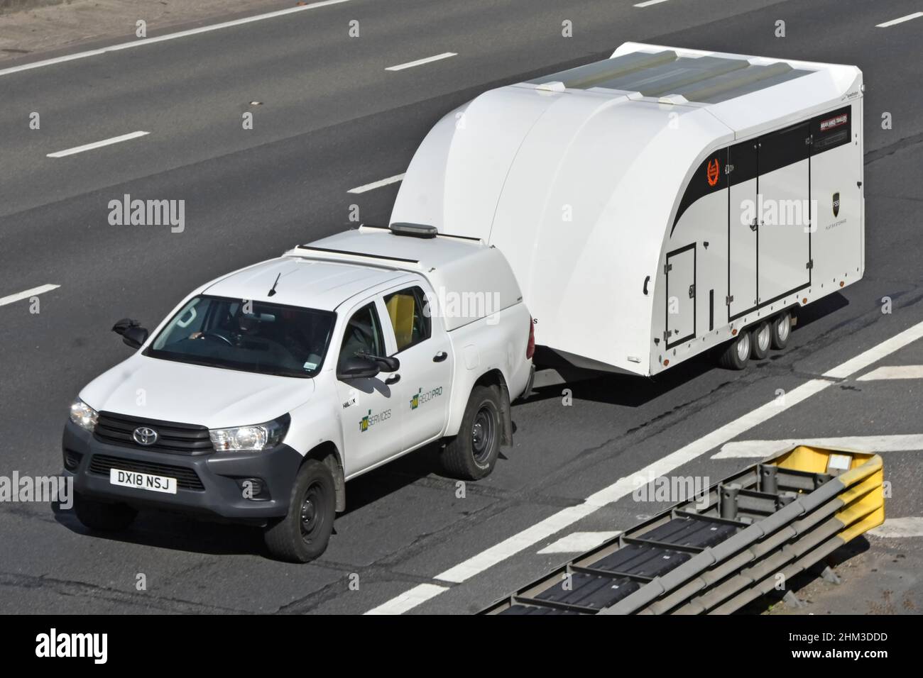 Vehicle delivery aerial side & front view Toyota Hilux pickup truck towing a white enclosed secure car transporter trailer driving on UK motorway road Stock Photo
