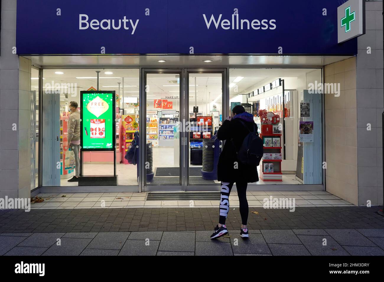 Boots Beauty Wellness store in shopping high street woman customer outside shopfront on pavement a dusk afternoon view at Christmas time England UK Stock Photo