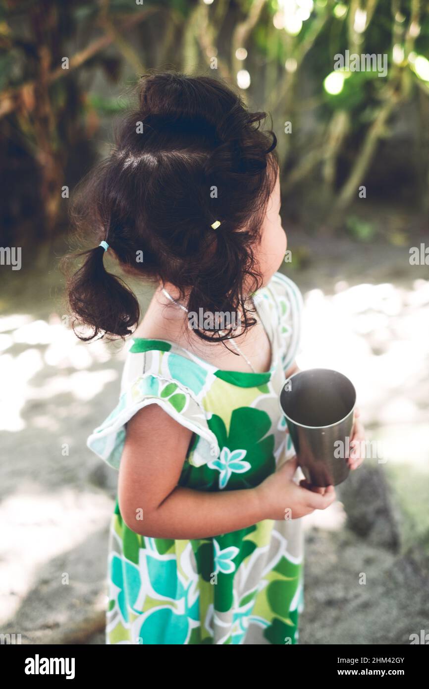 Unrecognizable cute little girl with curly hair looking faraway at the back holding stainless cup. Portrait orientation. Selected focus. Stock Photo