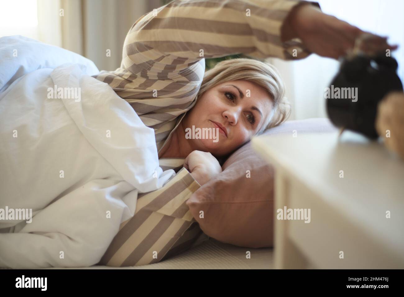 Overweight woman waking up in bed and switching off the alarm clock at home. Stock Photo