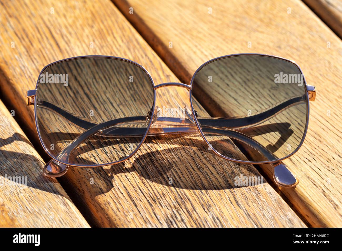 Metal thin trendy sunglasses dark tint, on wooden table. Sunny day, outdoors, bright light and shadow. Closeup image, minimal style. Stock Photo