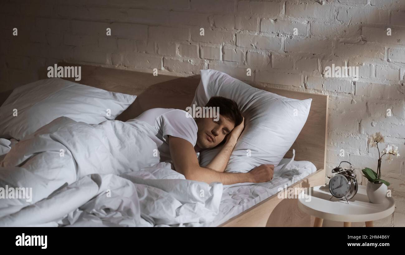 man sleeping in bed near alarm clock and potted orchid on bedside table Stock Photo