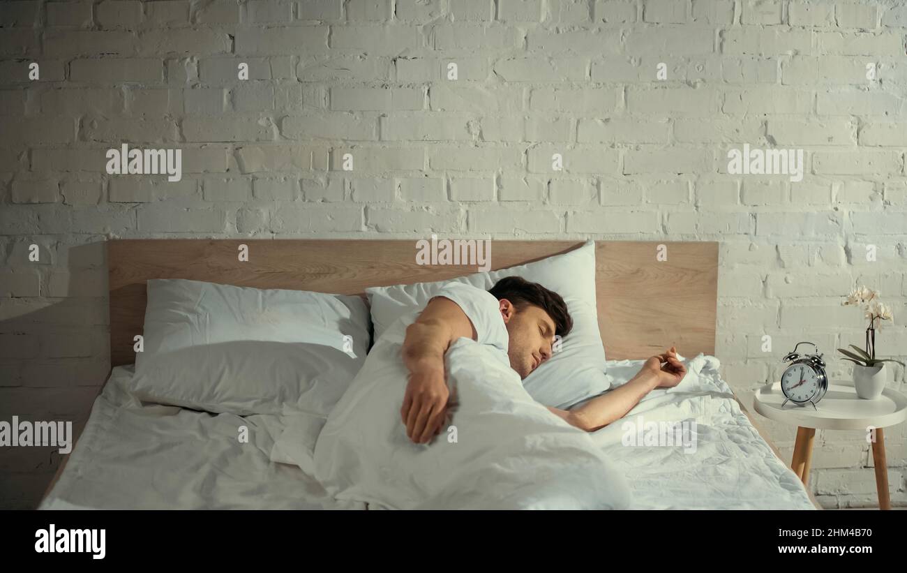 man sleeping on white bedding near vintage alarm clock and potted orchid on bedside table Stock Photo
