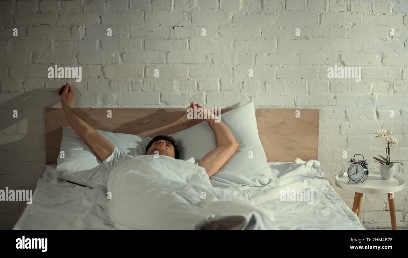 man stretching in bed near alarm clock and potted orchid on bedside table Stock Photo