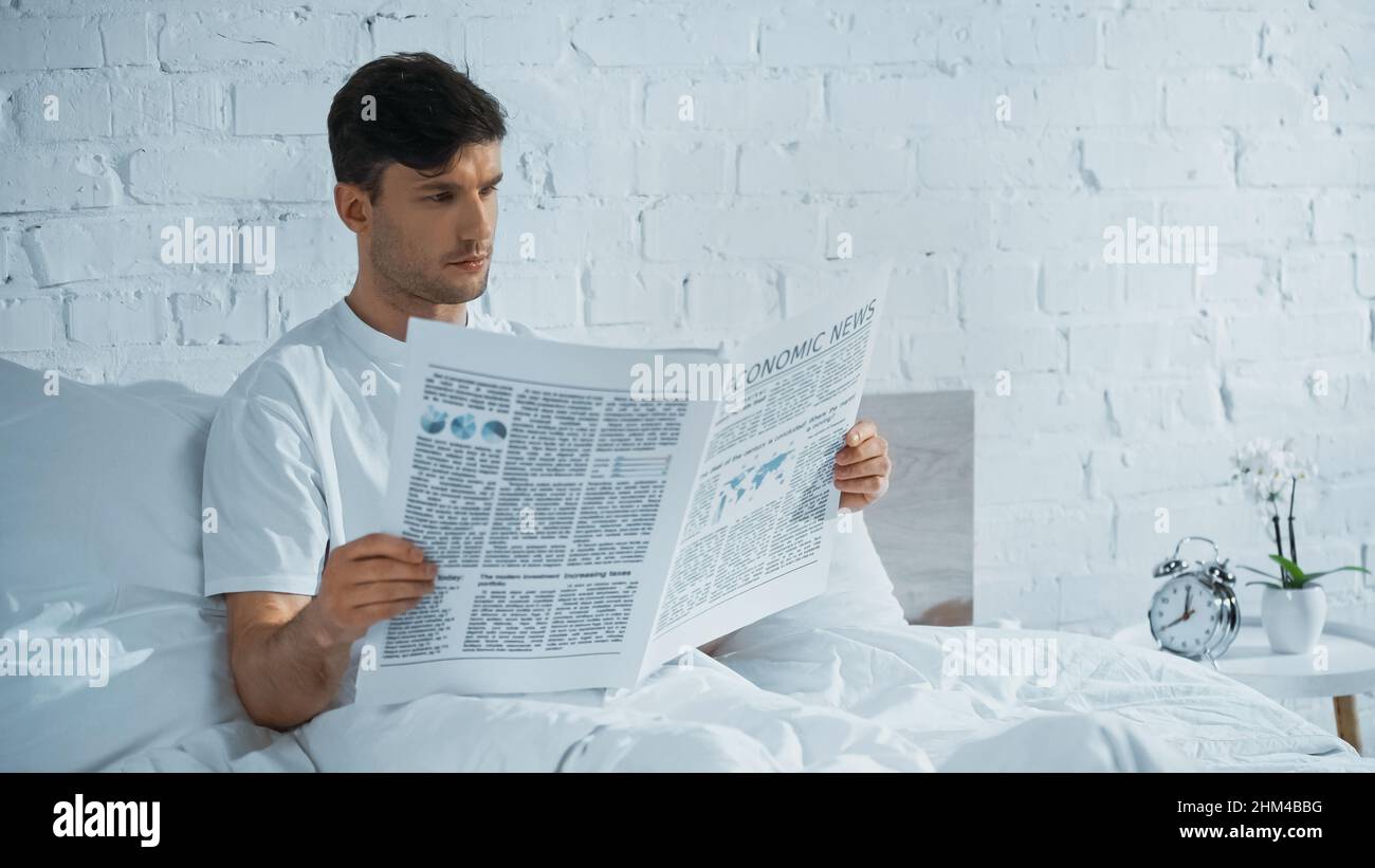 man reading economic news in bed near alarm clock and potted orchid on bedside table Stock Photo