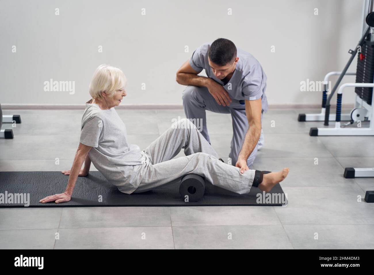 Aged lady sitting on mat with legs on roll Stock Photo