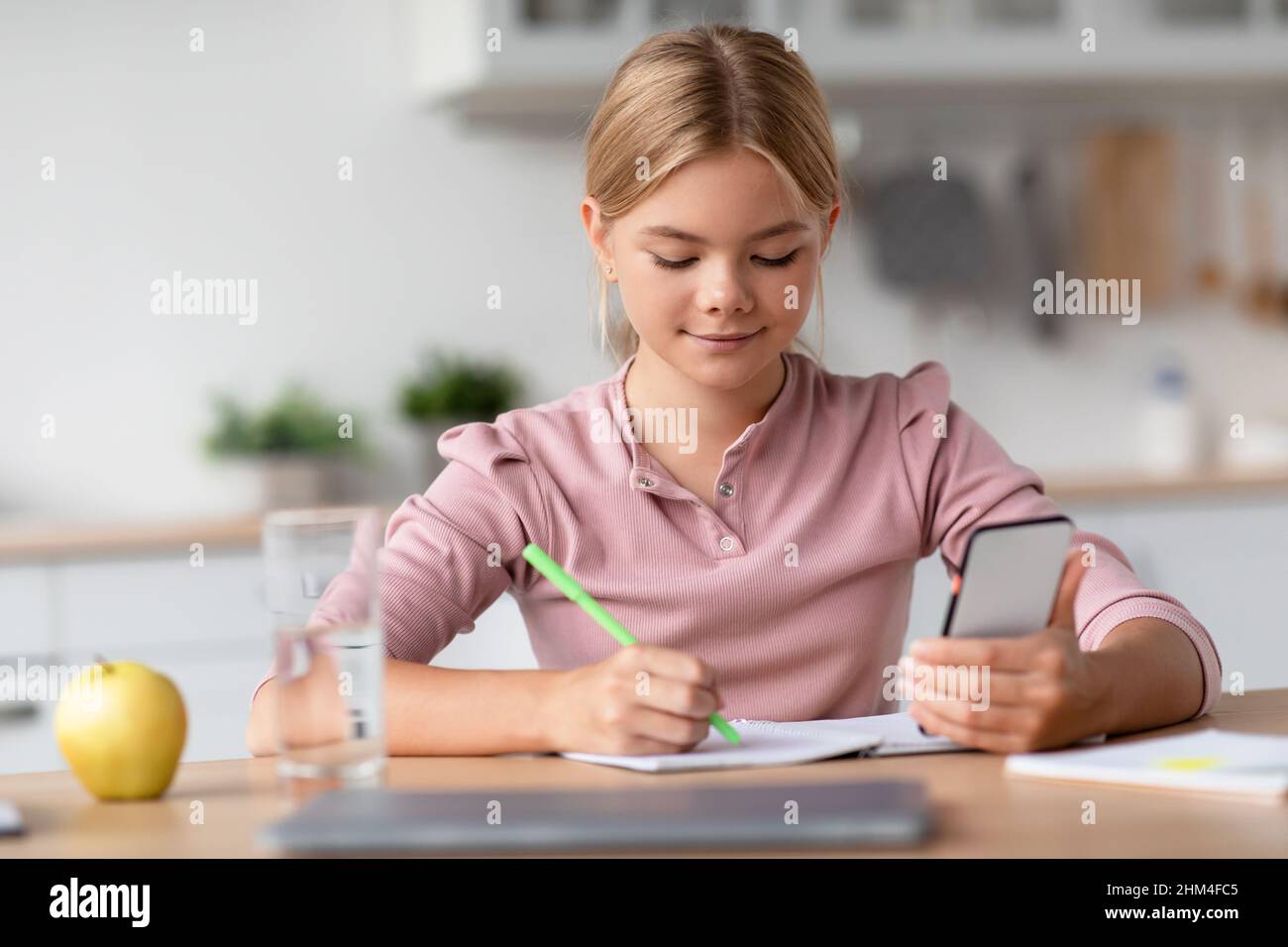 Serious pretty blonde european teenager girl makes notes with smartphone at table in minimalist kitchen Stock Photo